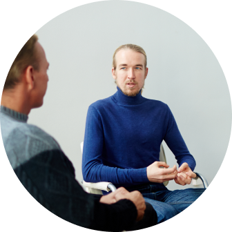 ADHD_sutrikimas_6 Two men sit facing each other in conversation; the man in a blue turtleneck gestures with his hands.