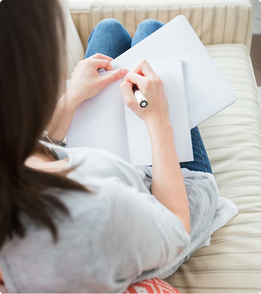ADHD_sutrikimas_8 Person sits on a sofa, writing in an open notebook with a pen at home.