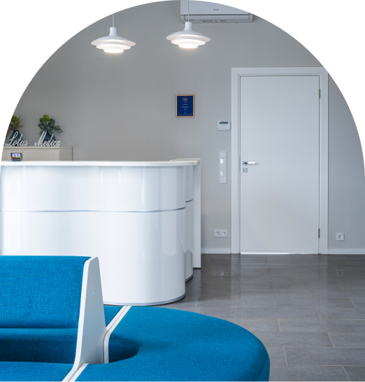 ADHD_sutrikimas_9 Modern clinic reception area with a curved white desk, blue seating in the foreground, gray tiled floor, and a closed white door in the back under two pendant lights.