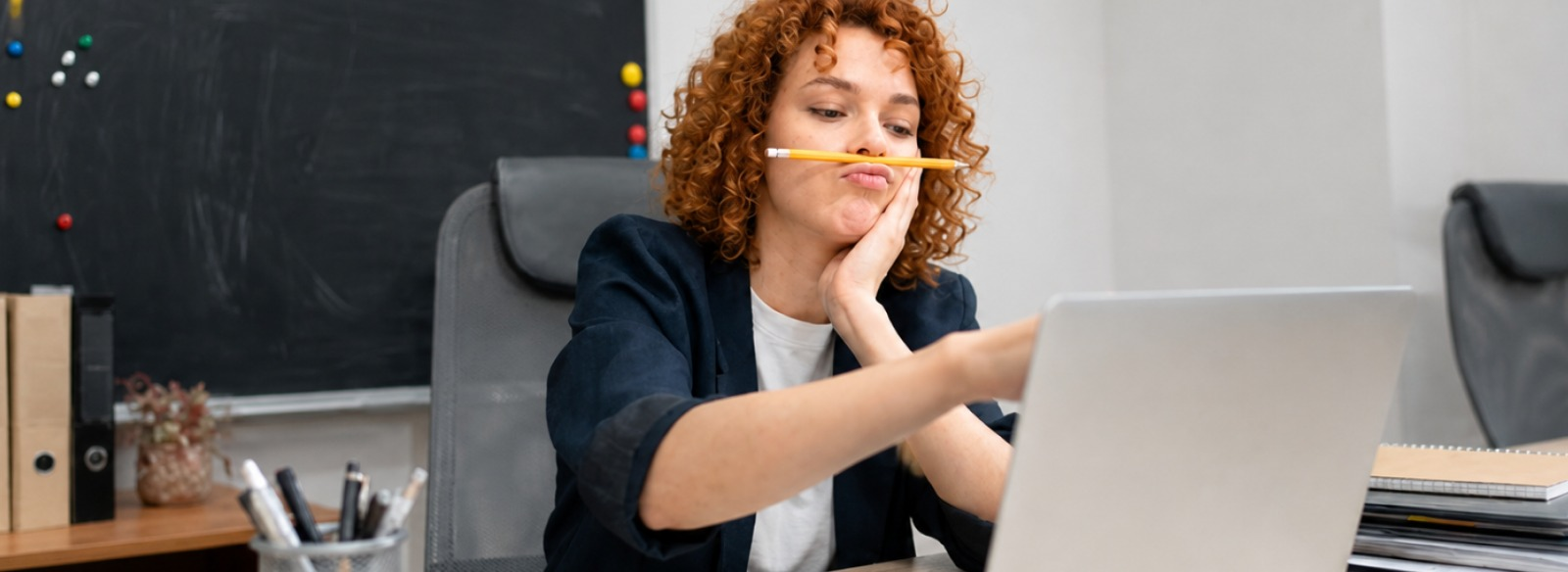 ADHD kas tai Woman with curly red hair sits at a desk with a pencil balanced between her lips, looking at a laptop screen.