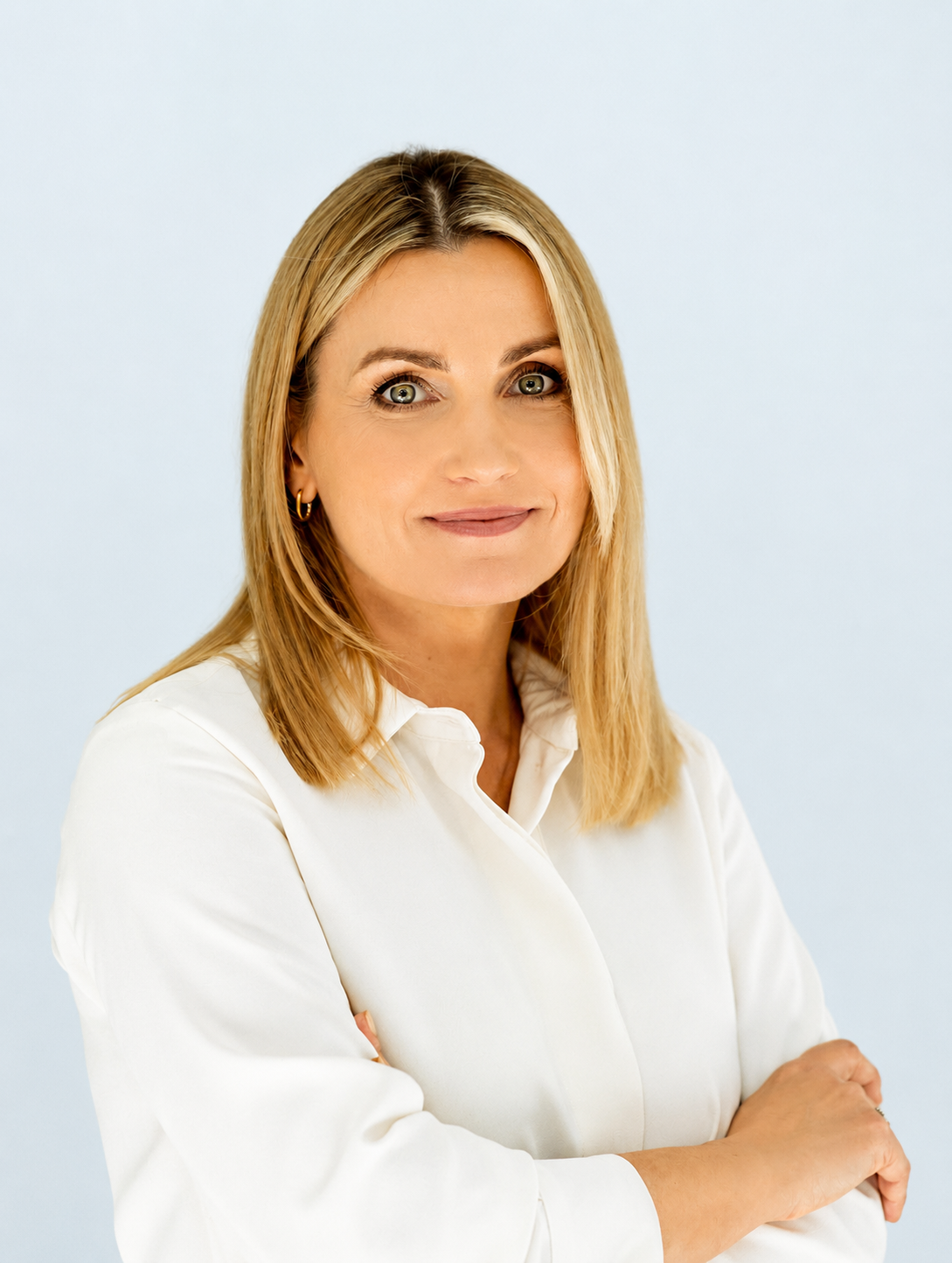 ChatGPT Image Jul 13, 2026, 11_25_46 AM Portrait of a smiling blonde woman in a white blouse with arms crossed against a light blue background