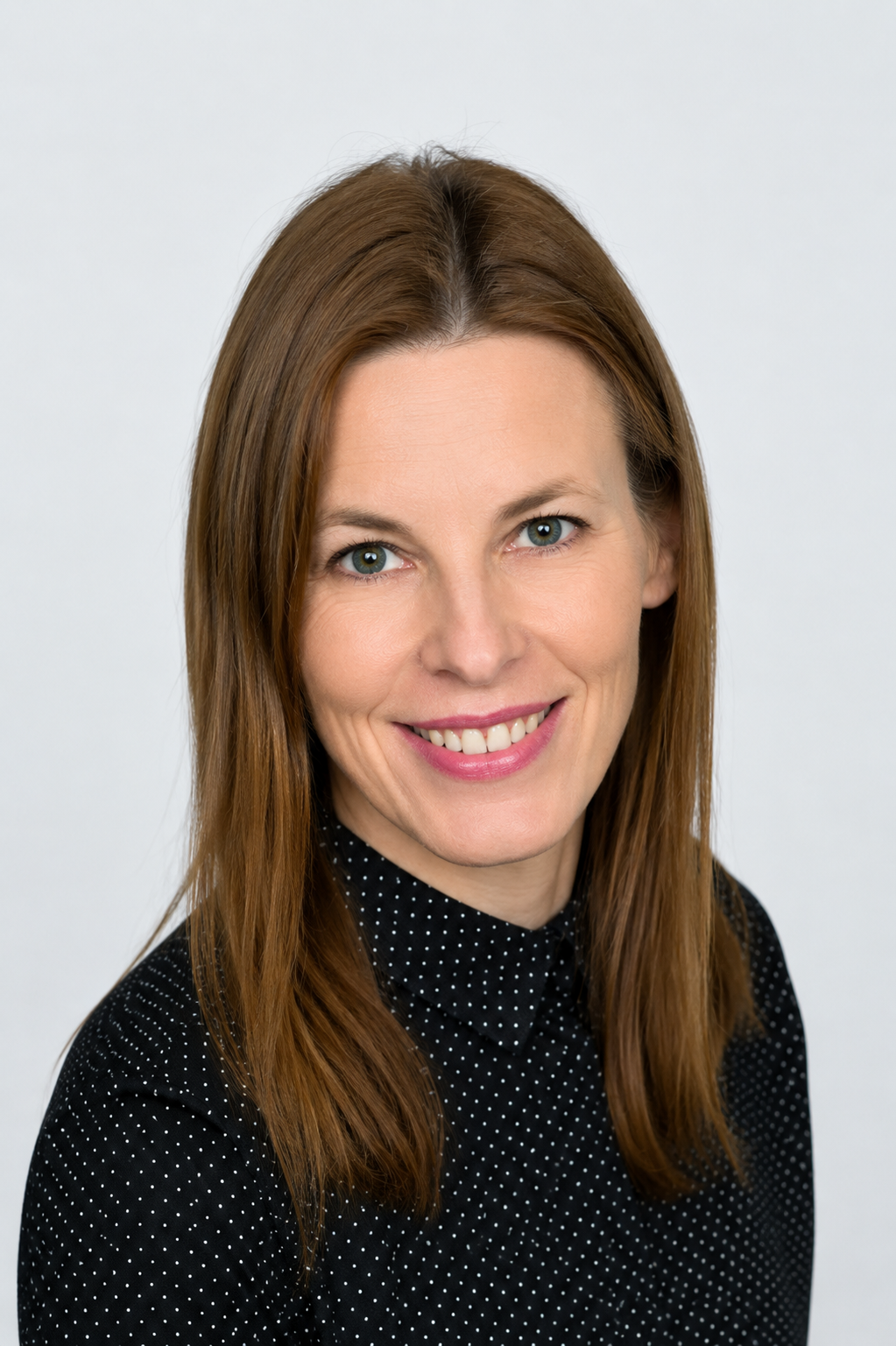 Head-and-shoulders portrait of a smiling woman with brown hair wearing a black polka-dot blouse against a light gray background.